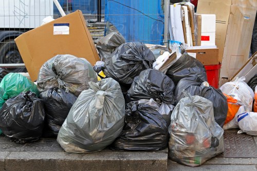 A collection of densely packed black and grey rubbish bags, made from plastic and filled with waste materials, is situated on a paved outdoor surface, likely on a street or alleyway. Some bags are tied at the top, and others appear loosely gathered, with a few showing partial contents through the thin plastic. The background features a cluttered environment with additional cardboard boxes, wooden pallets, and a red container, indicating a busy or industrial area, possibly near a waste collection or recycling point in Edmonton. The scene is illuminated by natural daylight, with the bags positioned in front of a blue structure or wall, suggesting an urban setting within the vicinity of Business Waste Removal Edmonton's service area. This visual highlights the typical accumulation of waste awaiting removal or processing, aligning with the company's rubbish removal services around the local town.