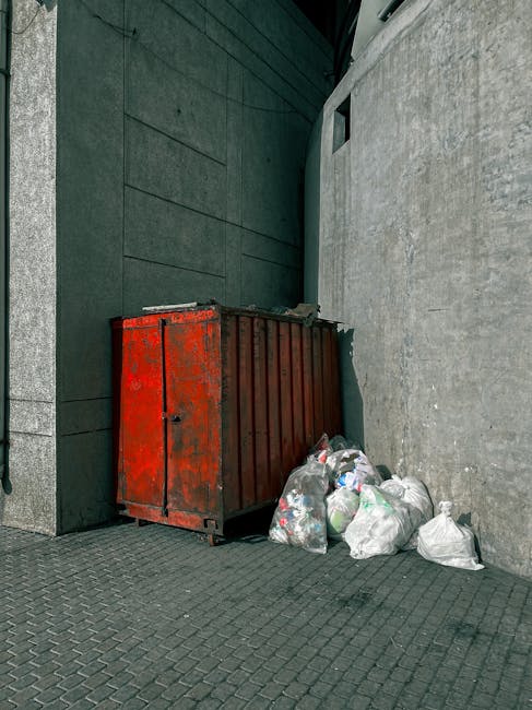 A large, industrial-style red metal waste container with a closed, slightly rusted and weathered surface, situated against a dark teal wall on a paved surface composed of small, black interlocking bri