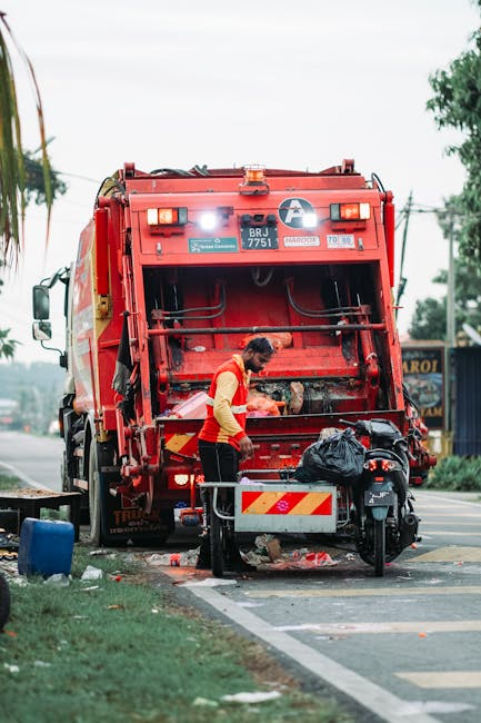 A waste collection worker wearing a high-visibility yellow vest with reflective stripes, operating a waste collection vehicle in an urban environment during evening hours, with buildings featuring ill
